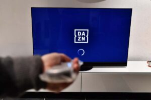 Una mano con telecomando in mano e una tv con logo Dazn sullo sfondo