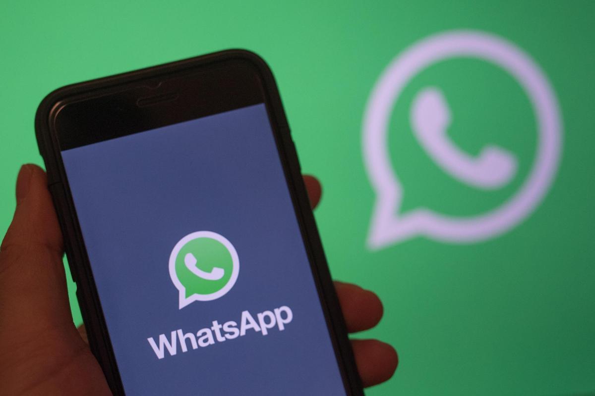 Schermata WhatsApp su uno smartphone tenuto in mano. Sullo sfondo logo WhatsApp